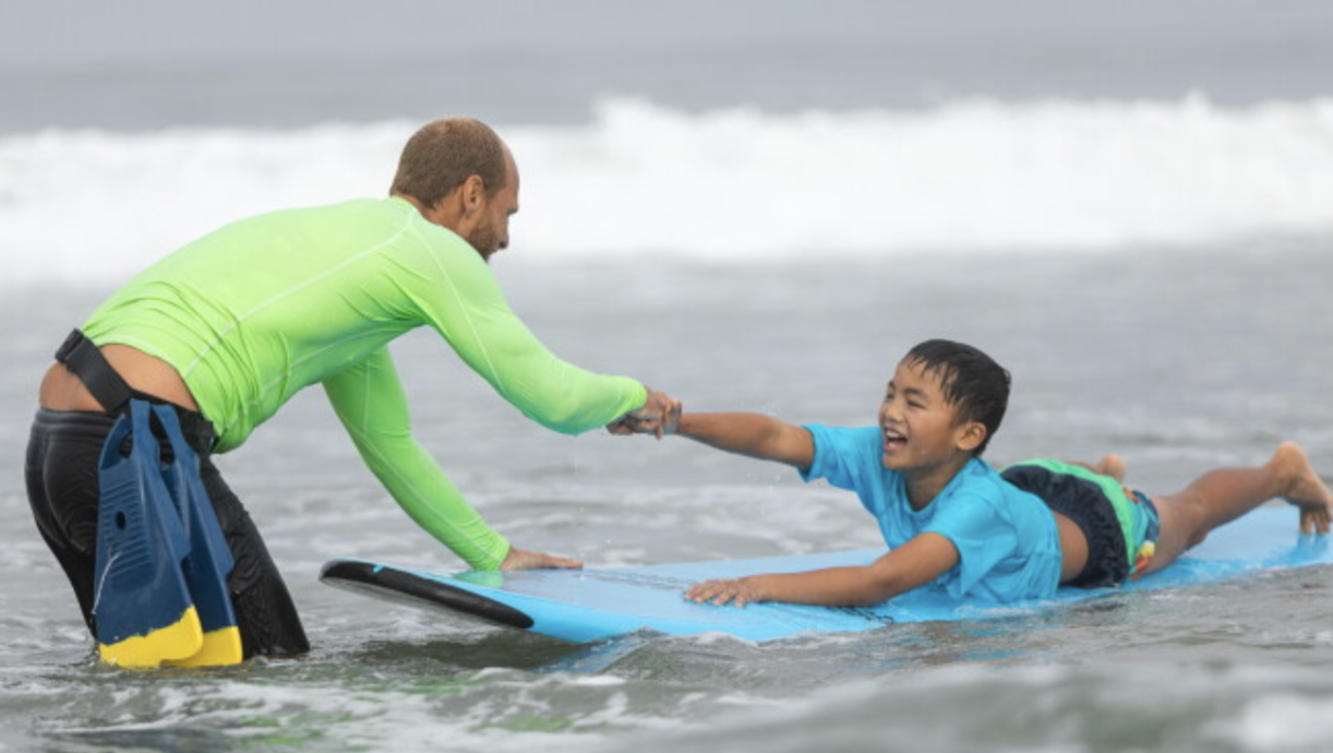 An Everybody Surf instructor and a smiling young adaptive surfer clasping hands on a surfboard in the water